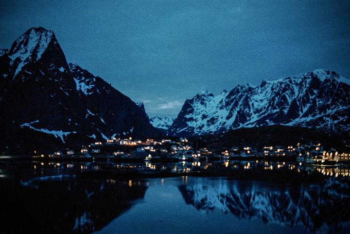 A small town with illuminated buildings sits at the base of snow-capped mountains, reflected in the still water of a lake at night.