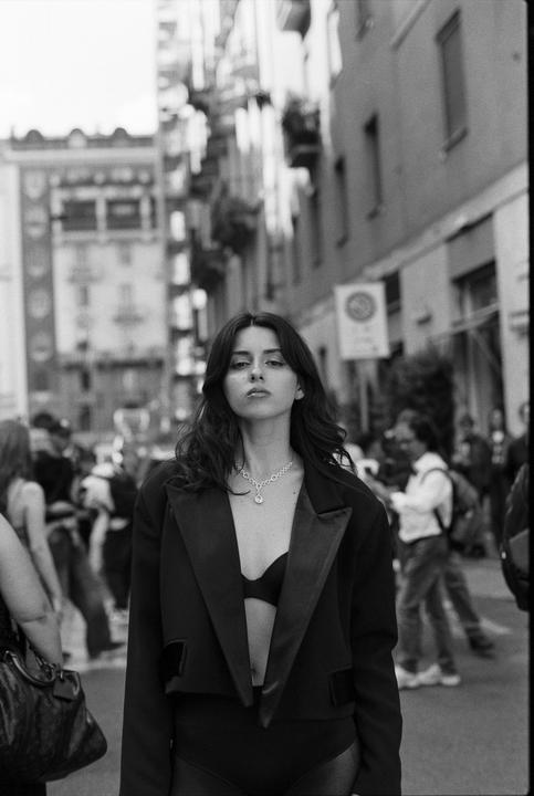 A woman wearing a blazer and bra stands on a city street with buildings and people in the background.
