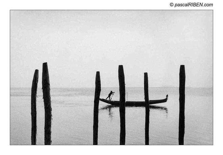 A man poles a gondola between wooden posts in the water.