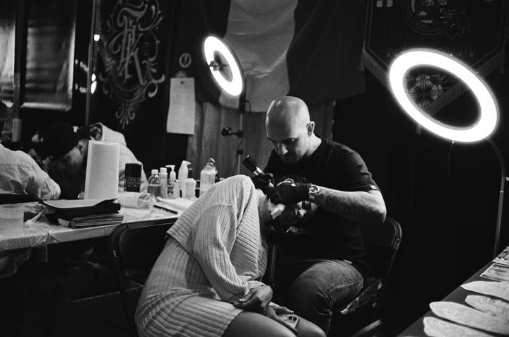 A man tattoos a woman's head while she sits with her head down, covered by a sweater.