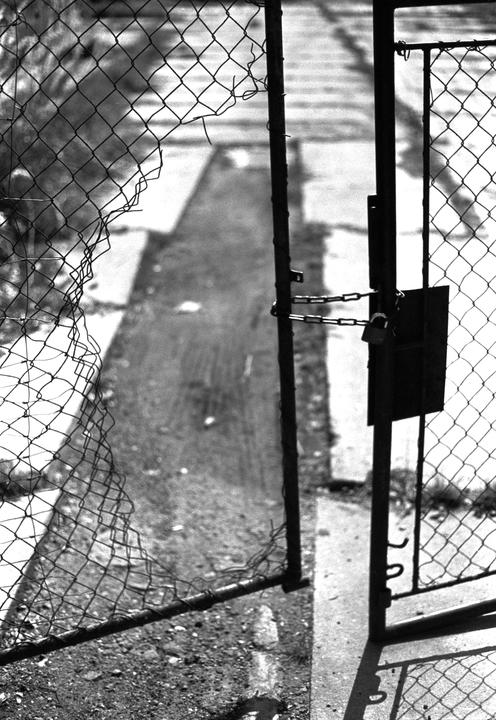 A black and white photo shows a broken chain link fence and a gate secured with a chain and padlock.