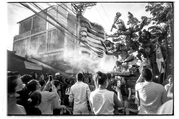 A crowd of people watches a large, ornate statue of mythical figures on a float, with smoke rising around it.