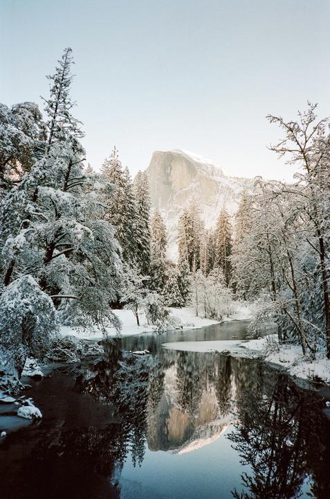 A snow-covered landscape features a river reflecting a large mountain and surrounding trees.