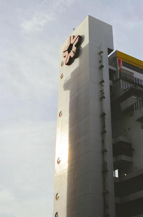 A tall, light-colored building with a large red symbol on its side is illuminated by sunlight, casting a long shadow.