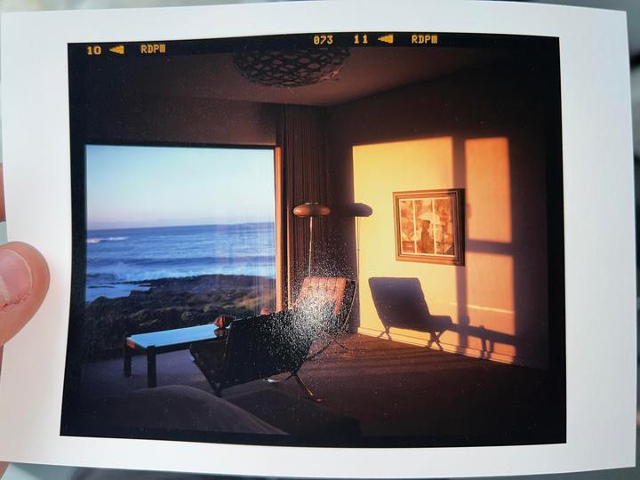 A room with two chairs and a table overlooks the ocean, with sunlight casting shadows on the wall.