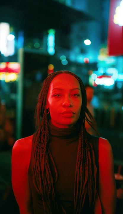 A woman with braided hair is illuminated by red and blue neon light at night.