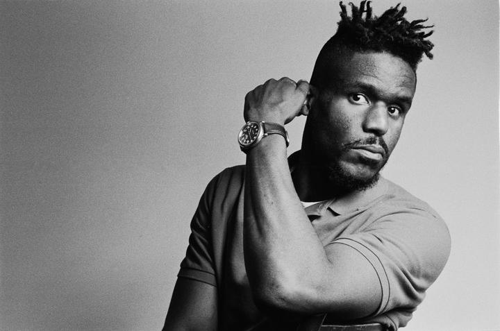A man with dreadlocks and a beard looks directly at the viewer, resting his arm on a chair.