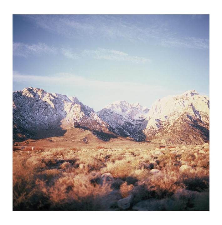 A vast desert landscape with dry brush and rocks in the foreground leads to snow-capped mountains under a clear sky.