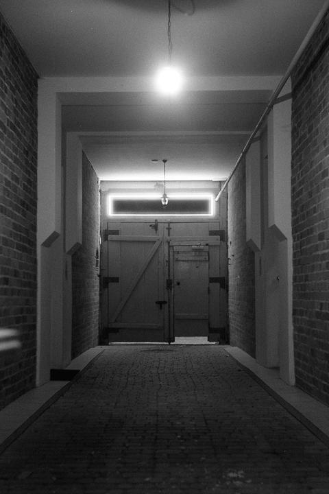A narrow, brick-lined alleyway leads to a large wooden door, illuminated by a single hanging lightbulb and a rectangular neon light.