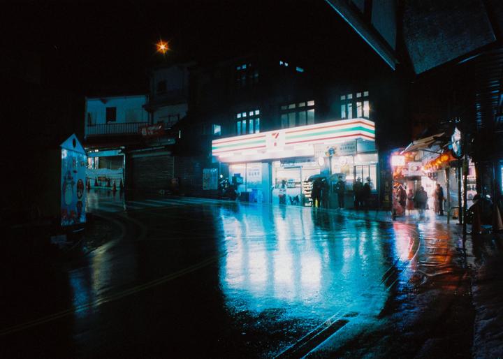 A brightly lit 7-Eleven store reflects on a wet street at night.