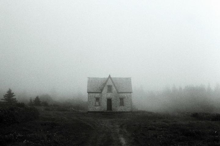 A small house with a peaked roof stands alone in a field on a foggy day.