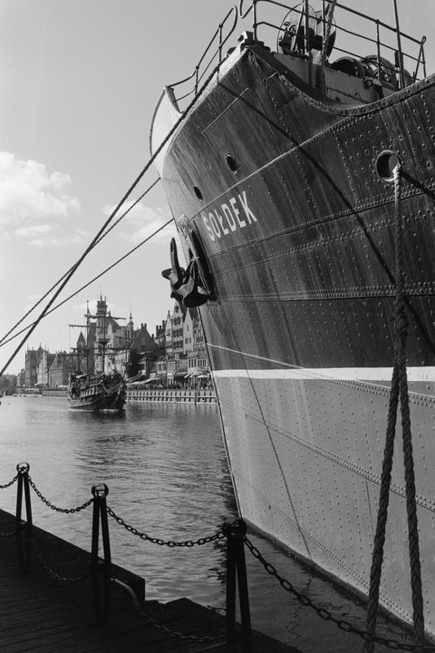 A large ship named Soldek is docked in a harbor with buildings and another ship in the background.