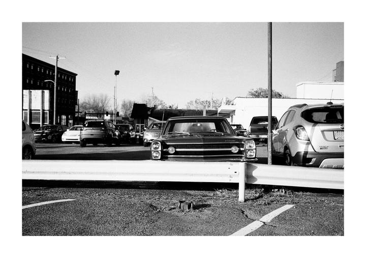 A black and white street photograph shows a vintage car parked in a lot behind a white barrier.