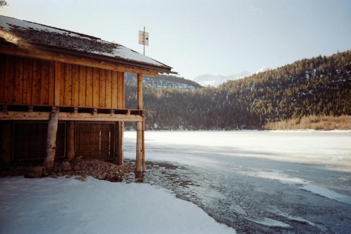 A wooden boathouse stands on the snowy shore of a frozen lake, with a forest and mountains in the background.