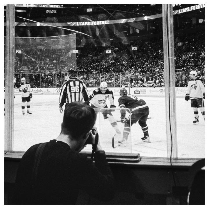 A man photographs a hockey game from behind the glass, with players and a referee on the ice and spectators in the stands.