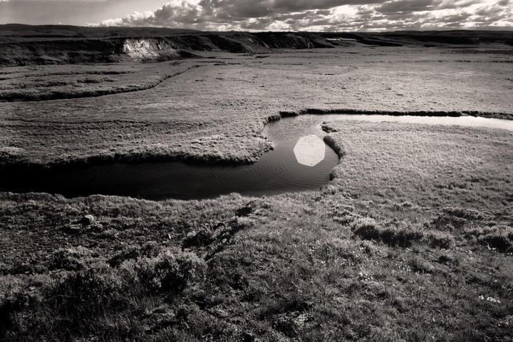 A river winds through a vast, grassy landscape under a cloudy sky.