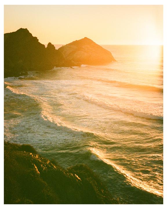 Waves crash against a rocky coastline as the sun sets over the ocean.