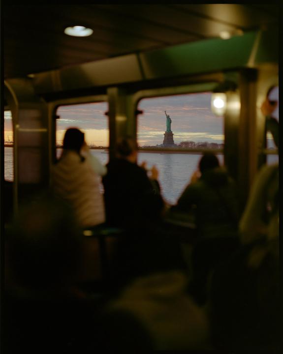 The Statue of Liberty is visible through a window from inside a ferry at sunset.
