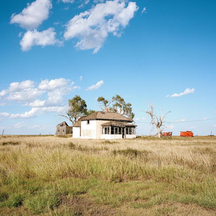 An abandoned house stands in a field of tall grass under a blue sky with scattered clouds.