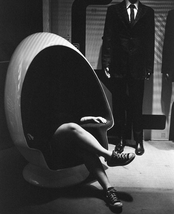 A person sits in a modern egg chair with their legs crossed, next to a mannequin dressed in a suit.