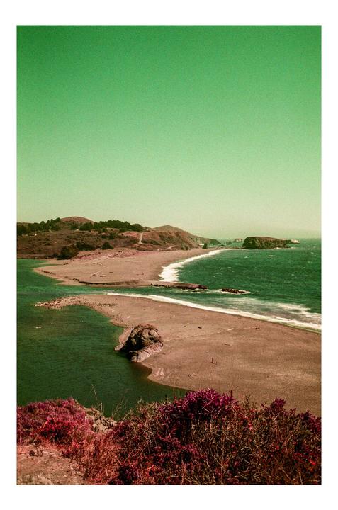A wide shot shows a beach with a river flowing into the ocean, with hills and an island in the background.