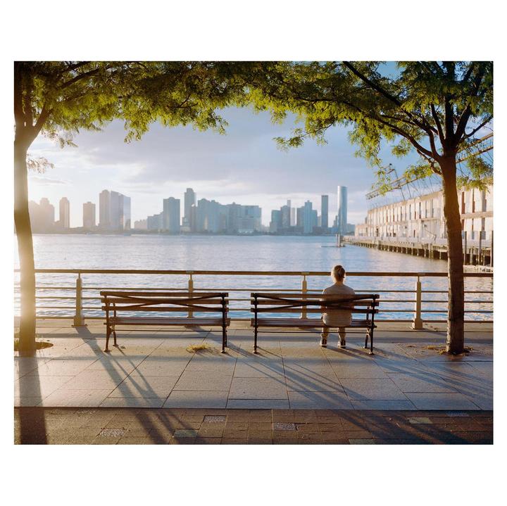 A person sits on a park bench overlooking the Hudson River with a city skyline in the distance.