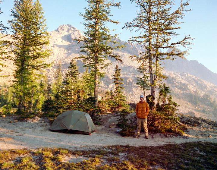 A man stands next to a tent in a mountainous landscape with pine trees.