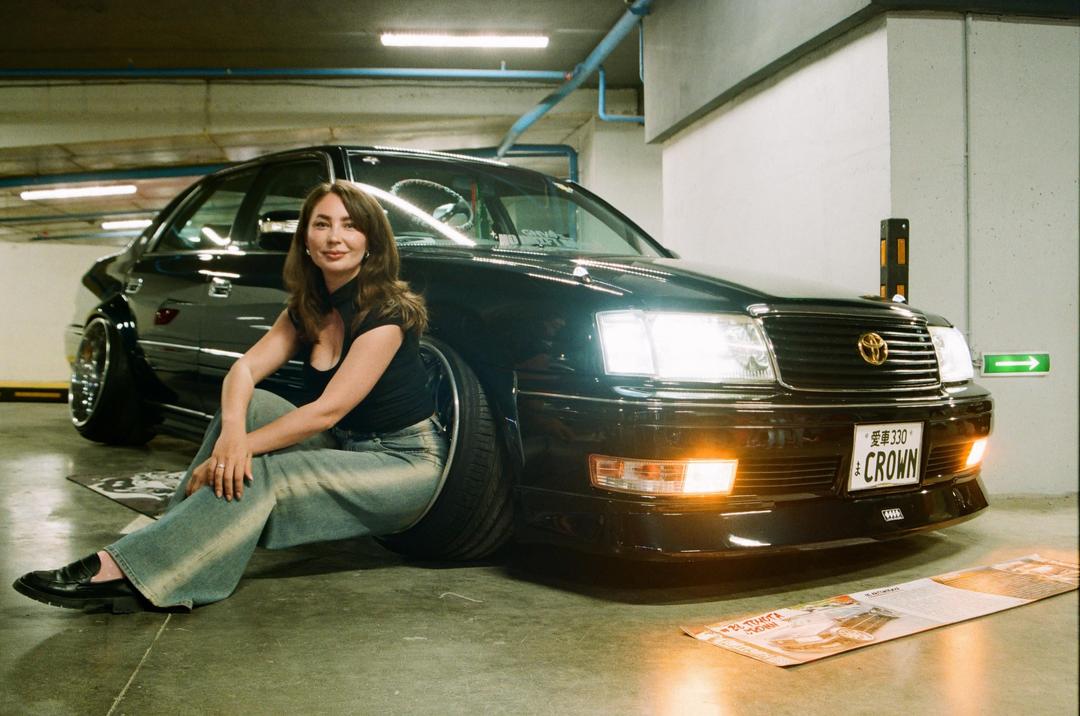 A woman sits on the ground next to a black car with its headlights on in a parking garage.