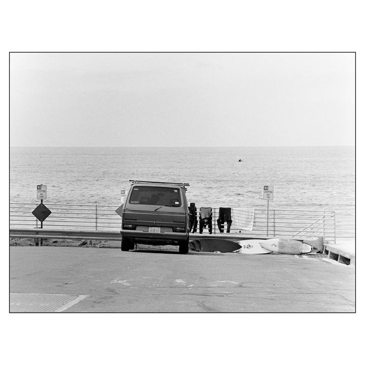 A van is parked next to a railing overlooking the ocean, with surfboards and wetsuits nearby.