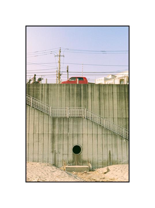 A red car is parked on a concrete wall with a staircase leading down to a sandy area.
