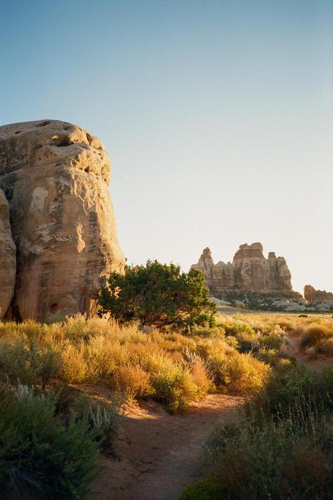 A dirt path leads through desert brush towards large rock formations under a clear sky.