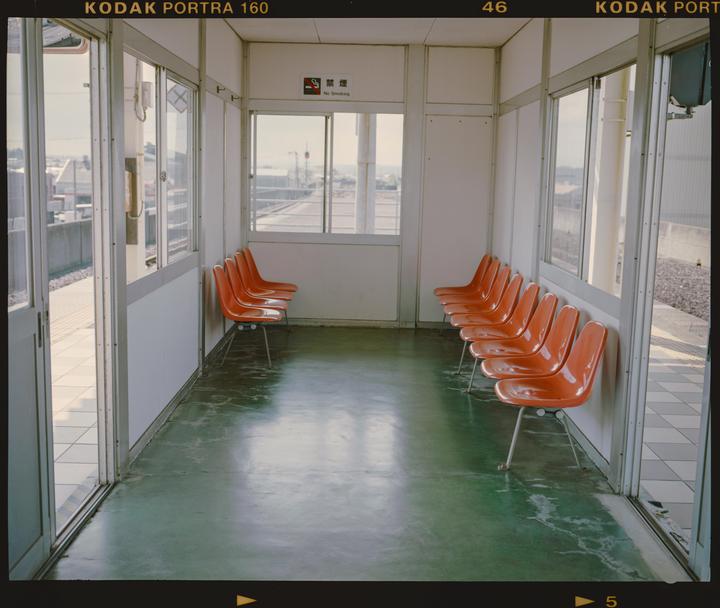 A row of orange chairs sits inside a waiting room with windows looking out onto train tracks.