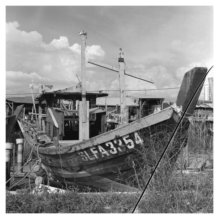 A weathered wooden fishing boat with the number "SLFA3354" painted on its bow sits on dry land amidst tall grass, with other structures and a cloudy sky in the background.