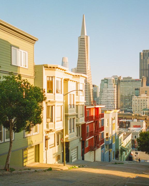 A street of colorful houses on a hill with a city skyline in the background during golden hour.