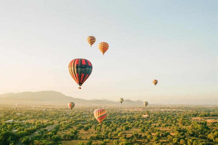 Several hot air balloons float above a green landscape with mountains in the distance.