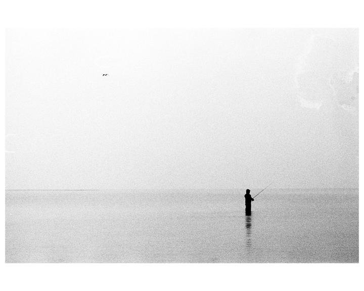 A lone fisherman stands in the water with a fishing rod under a hazy sky.
