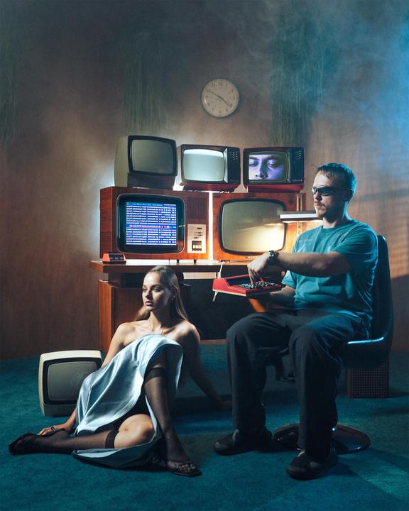 A man and a woman are in a room with multiple vintage televisions stacked on a wooden table.