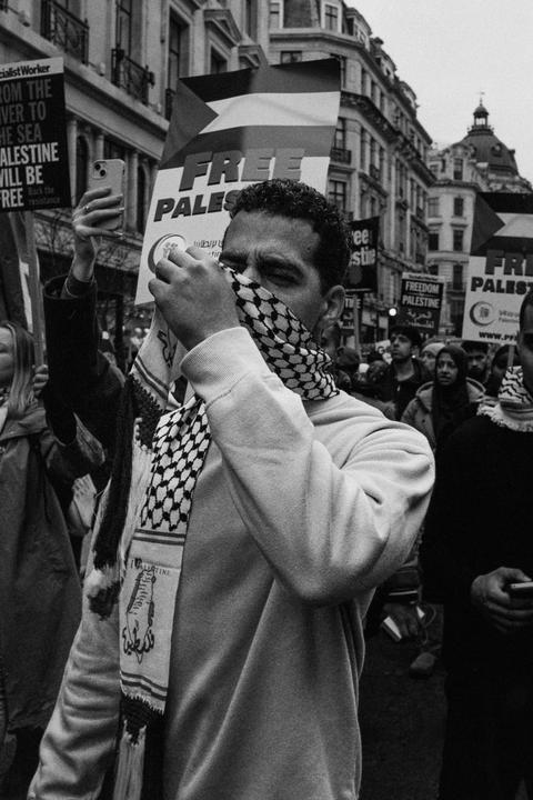 A man covers his face with a keffiyeh while holding a sign that reads "Free Palestine" at a protest.