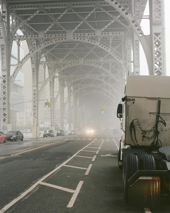A truck is parked on the side of a road under a large metal bridge on a foggy day.
