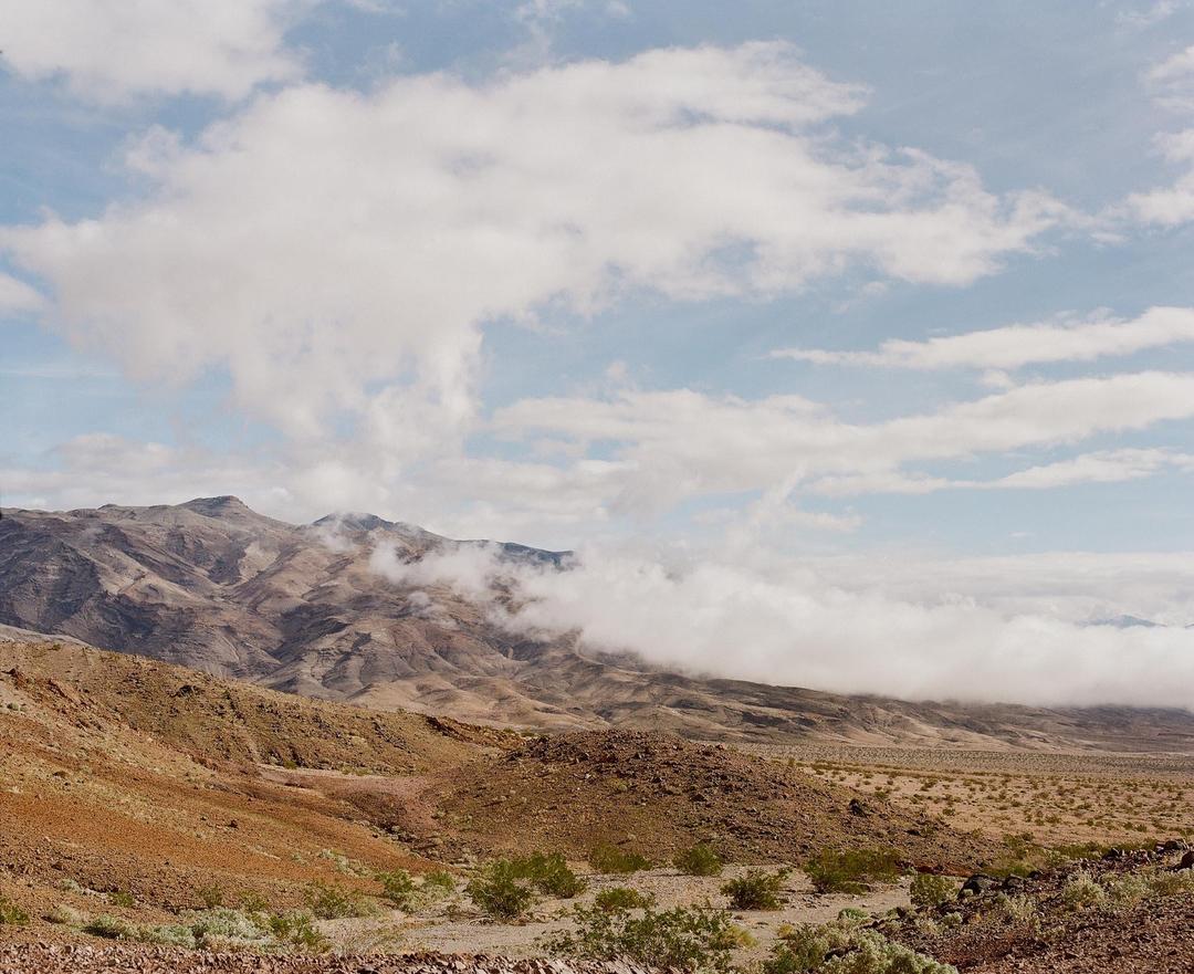 Clouds hang low over a desert mountain range with sparse vegetation in the foreground.
