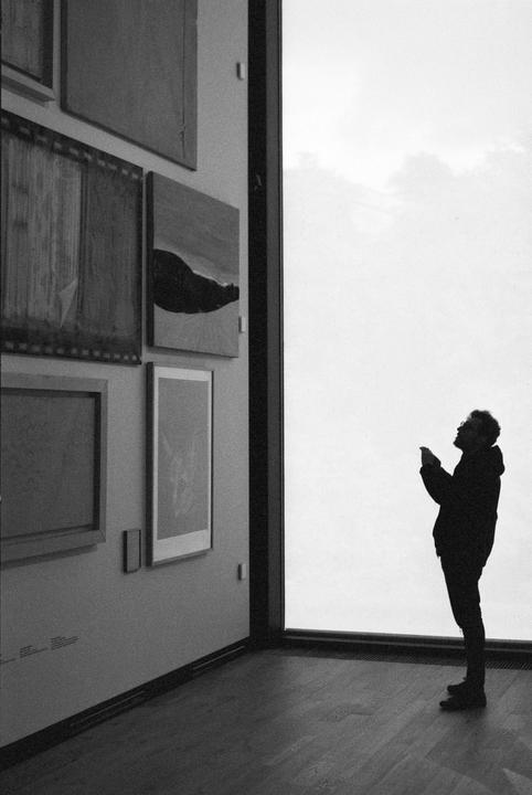 A man stands silhouetted against a bright window, looking up at something in his hands, with framed art on the wall to his left.