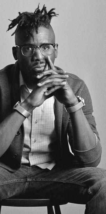 A man with dreadlocks and glasses sits with his hands clasped in front of him.