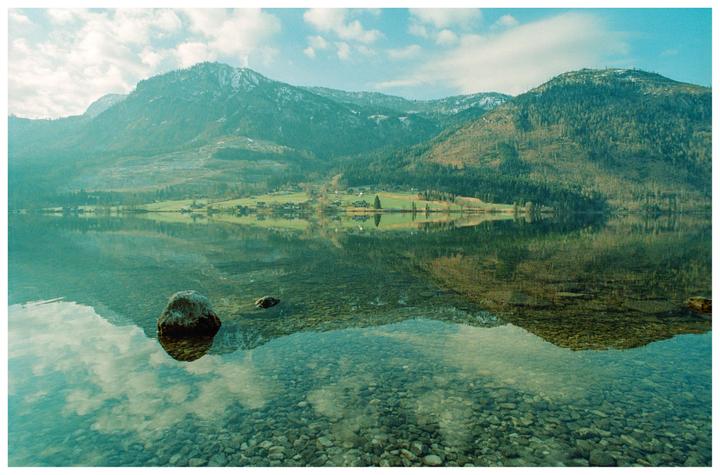 A calm lake reflects mountains and a village under a cloudy sky.