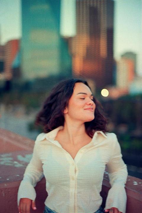 A woman with closed eyes and a smile leans against a railing with city buildings in the background.