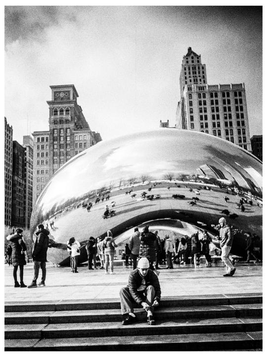 A man sits on steps in front of a large reflective bean-shaped sculpture with people and buildings visible in the background.