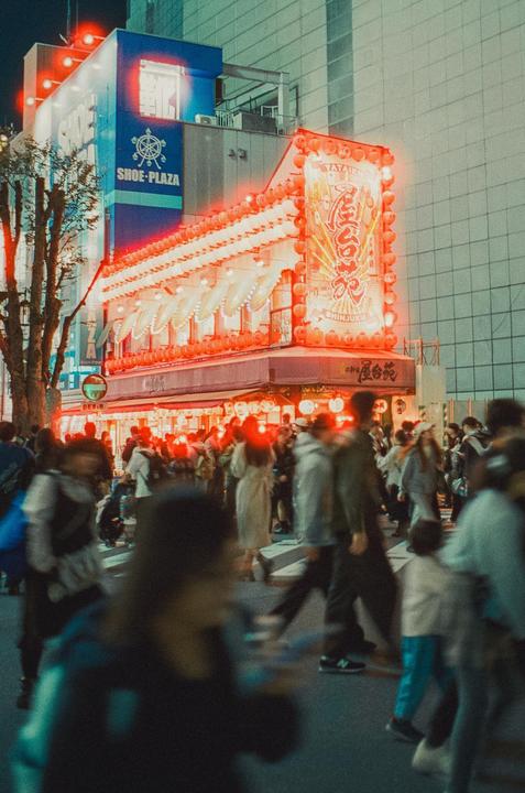A crowd of people walks past a brightly lit building with red lanterns and neon signs at night.