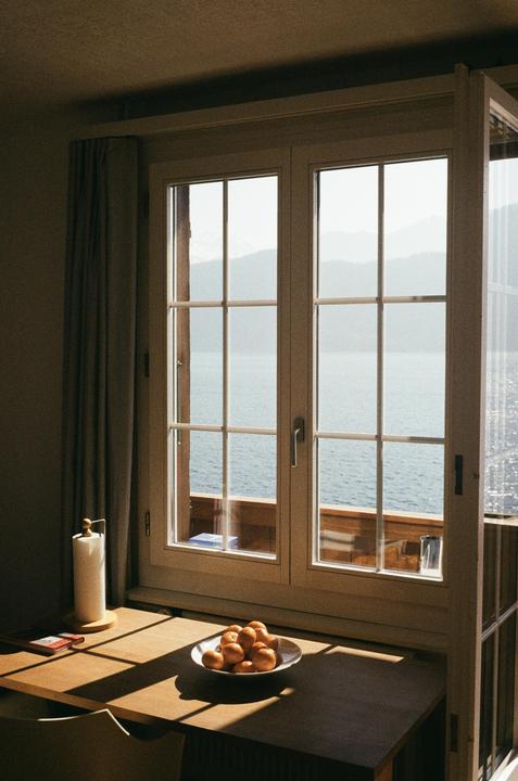A window overlooks a lake and mountains, with a bowl of oranges and a paper towel roll on a sunlit wooden table.
