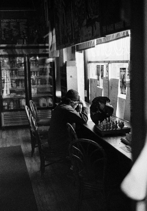 A person sits at a counter with a chess set, looking out a window.