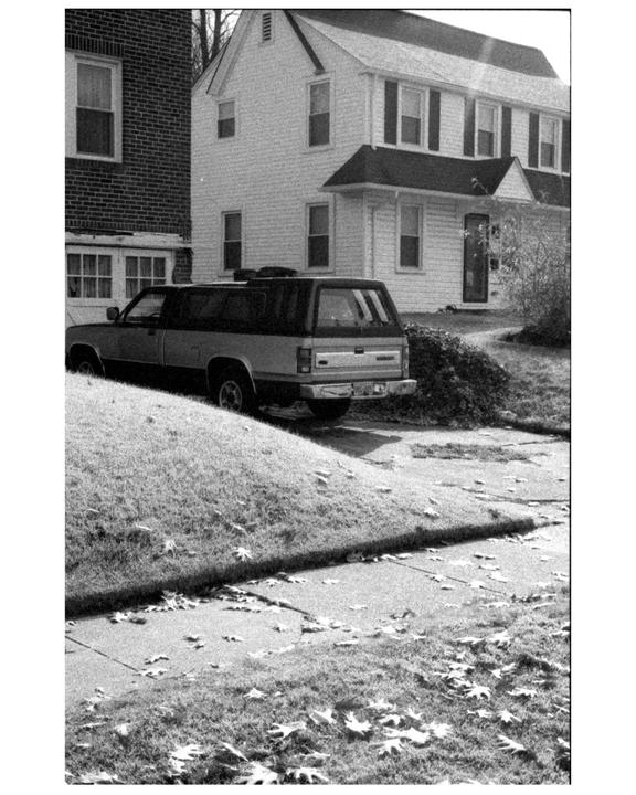 A black and white photograph shows a suburban street with a car parked in a driveway next to two houses.