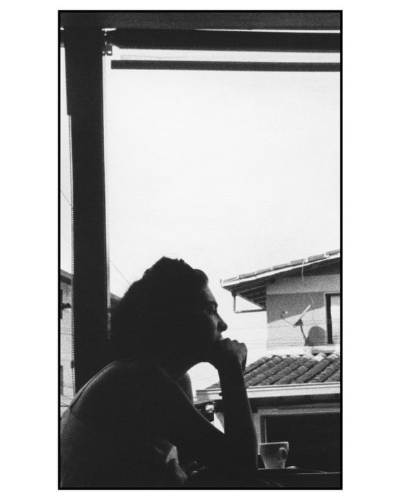 A woman in silhouette looks out a window with a cup of coffee in front of her.